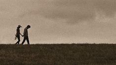 two people walking across a grass covered field under a cloudy sky with a kite in the air