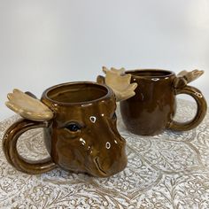 two coffee mugs with moose heads on them sitting on a tableclothed surface