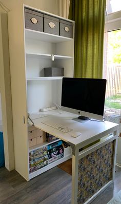 a desk with a computer on top of it next to a book shelf and window