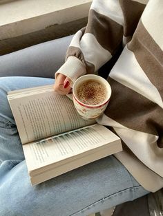 a person sitting on a couch reading a book and holding a cup of hot chocolate