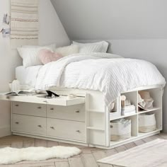 a white bed sitting under a slanted ceiling next to a dresser and shelf filled with books