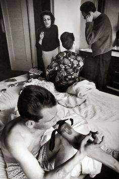 three people standing and sitting around a bed in a room with one person laying on the bed