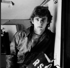 black and white photograph of a young man in leather jacket leaning against wall holding books