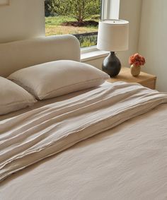 a bed with white sheets and pillows next to a lamp on a table in front of a window