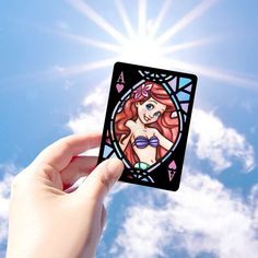 a hand holding up a playing card in front of blue sky with clouds and sun