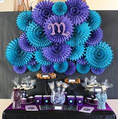 a purple and blue dessert table set up with paper fans on the wall behind it