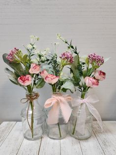 three glass vases with flowers in them on a white wooden table and one has a pink ribbon tied around it