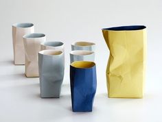 five different colored vases sitting next to each other on a white surface with one yellow cup in the middle