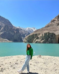 a woman standing on top of a sandy beach next to a mountain lake with snow capped mountains in the background