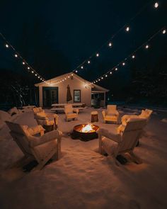 a fire pit in the middle of a snow covered yard with chairs and lights strung over it