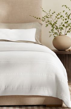 a bed with white sheets and pillows on top of it next to a plant in a vase