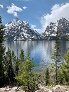 the mountains are covered in snow and some trees by the water with blue sky above them