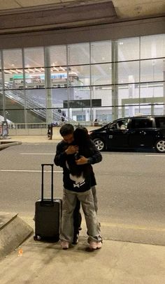 a man and woman hugging on the sidewalk with luggage in front of an airport terminal