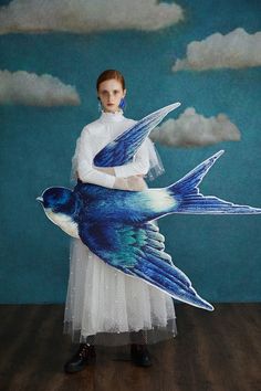 a woman in a white dress holding a blue bird with clouds on it's back