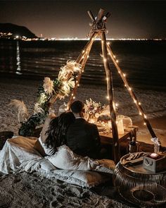 a man and woman are sitting in front of a teepee on the beach at night