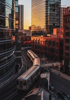 a train traveling down tracks next to tall buildings in a city at sunset with the sun going down