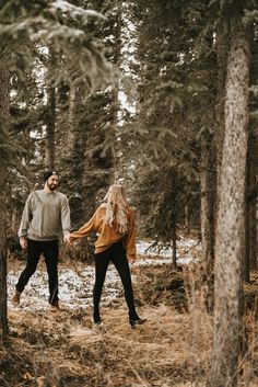 a man and woman holding hands walking through the woods in front of snow covered trees