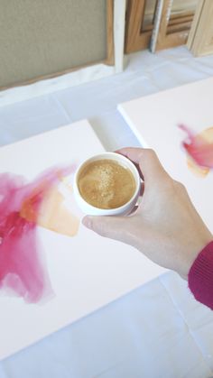 a person holding a cup of coffee in front of some paintings on the floor and table