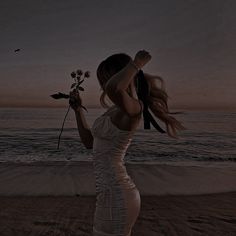 a woman standing on top of a sandy beach next to the ocean holding a flower