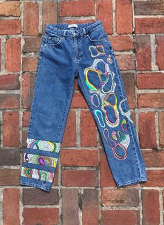 a pair of blue jeans with an embroidered design on the bottom and side, sitting against a brick wall