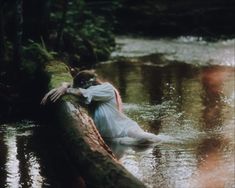 a woman in a white dress is laying on a log over a river with water flowing behind her