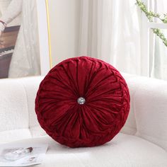 a red round pillow sitting on top of a white couch next to a book and plant