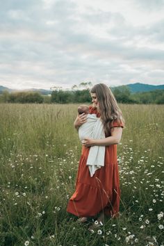 a woman in an orange dress holding a baby while standing in a field with daisies