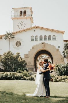 a bride and groom standing in front of a building with a clock on the tower