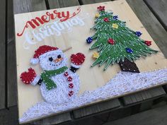 a christmas card with a snowman and a pine tree on it, sitting on top of a wooden board