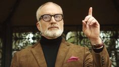 an older man with glasses pointing to something in the air while wearing a brown suit and black turtle neck sweater