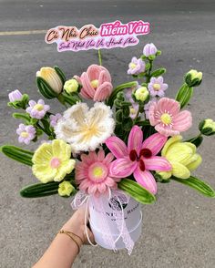 a hand holding a vase filled with flowers on the side of a road next to a street