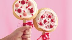 a hand holding two decorated cookies and lollipops on a stick with pink background