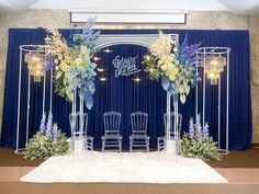 a stage set up with blue curtains and white chairs, decorated with floral arrangements on the floor