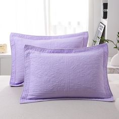 two purple pillows sitting on top of a bed next to a vase filled with flowers