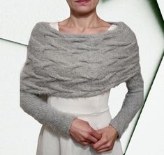 a woman standing in front of a white wall wearing a gray knitted shawl