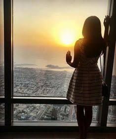 a woman standing in front of a window looking out at the city and sunsets