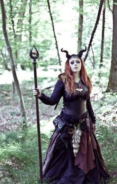 a woman dressed up as a demon holding a staff in the middle of a forest