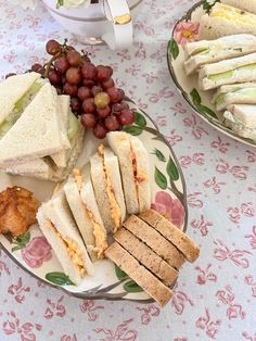 two plates with sandwiches, grapes and crackers on them sitting on a floral table cloth