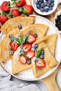 crepes with berries and powdered sugar are on a plate next to bowls of fruit