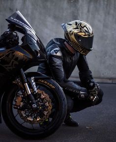 a person wearing a helmet and gloves sitting on a black motorcycle with gold accents, leaning against a wall