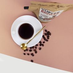 a cup of coffee next to beans and a bag of coffee on a pink surface