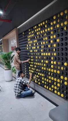 two men are sitting on the ground near a wall with yellow and black bottles attached to it