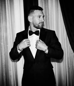 a man in a tuxedo adjusts his bow tie while standing next to curtains