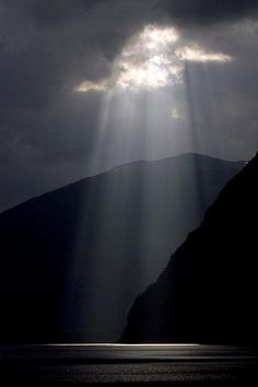 the sun shines through clouds over mountains on a dark, cloudy day at sea