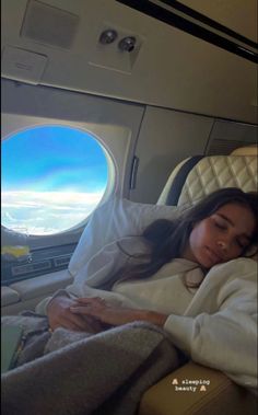 a woman laying in an airplane bed under a blanket and looking out the window at the sky