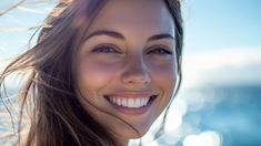 a close up of a woman smiling with her hair blowing in the wind and water behind her