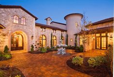 a large house with a fountain in the front yard at night time, surrounded by greenery and landscaping