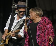 a man and woman singing into microphones at an outdoor music festival with one holding a guitar