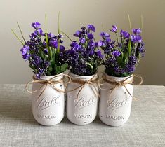 three white mason jars with purple flowers tied around the top and bottom, sitting on a table