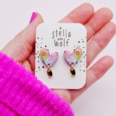 a hand holding a pair of pink and yellow heart shaped earrings with black stones on them